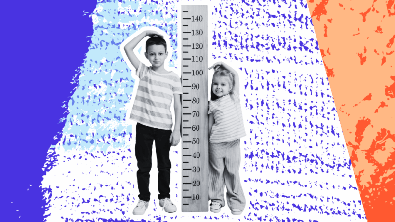 Two children stand next to a large height chart; the older child measures his height with his hand while the younger child smiles, highlighting differences in size.
